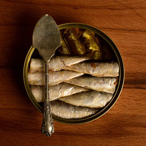 A round tin of small sardines. Some sardines have been removed from the top layer, revealing the lower layer in olive oil. There is a spoon balanced on the tin.