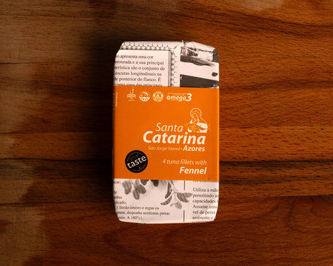 A tin of Santa Catarina tuna in newspaper-style packaging with an orange label against a wood background