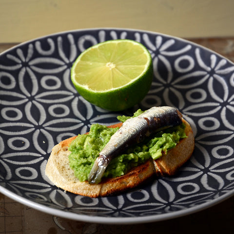 Mashed avocado on a slice of toasted bread topped with a sardine, on a patterned plate.