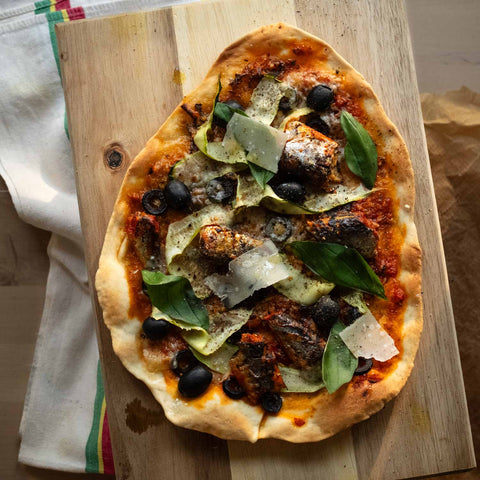 Tomato passata, ribbons of courgette, smoked sardines, sliced mozzarella, salt & pepper, torn basil leaves, all on top of pizza dough, placed on a wooden chopping board.
