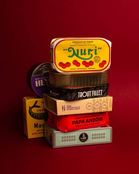 A selection of tinned fish stacked on top of each other, displayed against a dark red background showcasing notable brands including Nuri, Papa Anzois and Real Conservera.