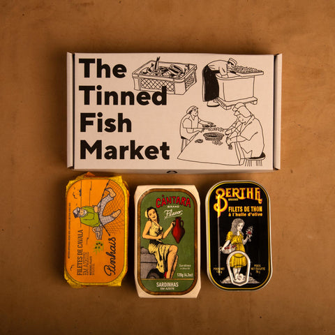 Three colourfully packaged tins of fish beneath a gift box illustrated with line drawings of the cannery workers, a basket of fish and The Tinned Fish Market in bold black lettering.