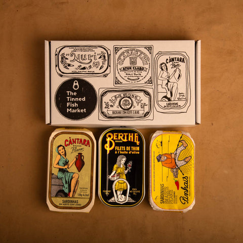 Three colourfully packaged tins of fish beneath a white cardboard gift box illustrated with black line drawings of the tins and The Tinned Fish Market logo.