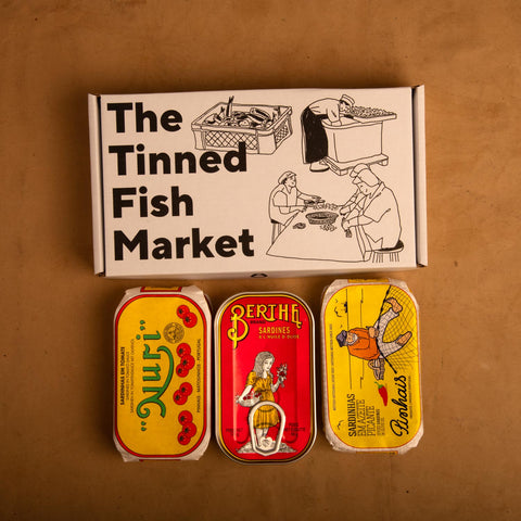 Three colourfully packaged tins of fish beneath a gift box illustrated with line drawings of the cannery workers, a basket of fish and The Tinned Fish Market in bold black lettering.