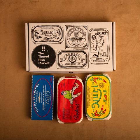 Three colourfully packaged tins of fish beneath a gift box illustrated with line drawings of the tins and The Tinned Fish Market logo.
