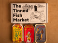 Three colourfully packaged tins of fish beneath a gift box illustrated with line drawings of the cannery workers, a basket of fish and The Tinned Fish Market in bold black lettering.