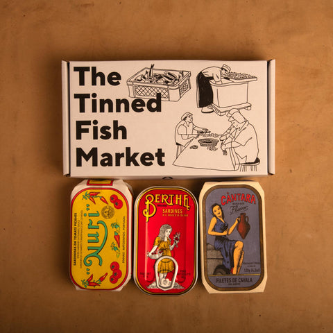 Three colourfully packaged tins of fish beneath a gift box illustrated with line drawings of the cannery workers, a basket of fish and The Tinned Fish Market in bold black lettering.