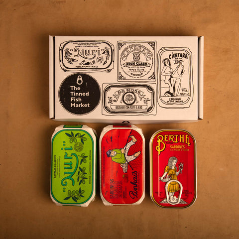 Three colourfully packaged tins of fish beneath a gift box illustrated with line drawings of the tins and The Tinned Fish Market logo.