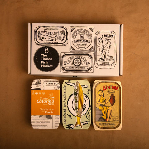 Three colourfully packaged tins of fish beneath a gift box illustrated with line drawings of the tins and The Tinned Fish Market logo.