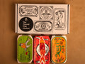 Three colourfully packaged tins of fish beneath a gift box illustrated with line drawings of the tins and The Tinned Fish Market logo.