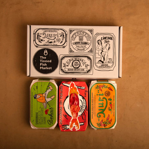 Three colourfully packaged tins of fish beneath a gift box illustrated with line drawings of the tins and The Tinned Fish Market logo.