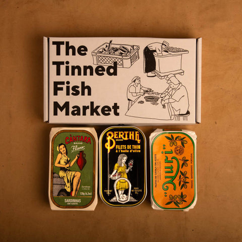 Three colourful tins of Portuguese tinned fish above a cardboard gift box, against a light brown background.