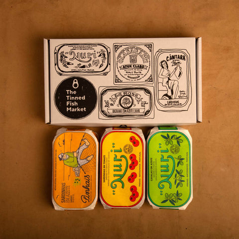 Three colourfully packaged tins of fish beneath a white cardboard gift box illustrated with black line drawings of the tins and The Tinned Fish Market logo.
