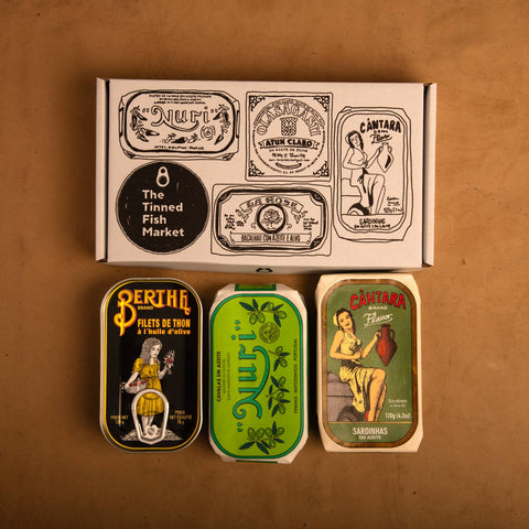 Three colourfully packaged tins of fish beneath a white cardboard gift box illustrated with black line drawings of the tins and The Tinned Fish Market logo.
