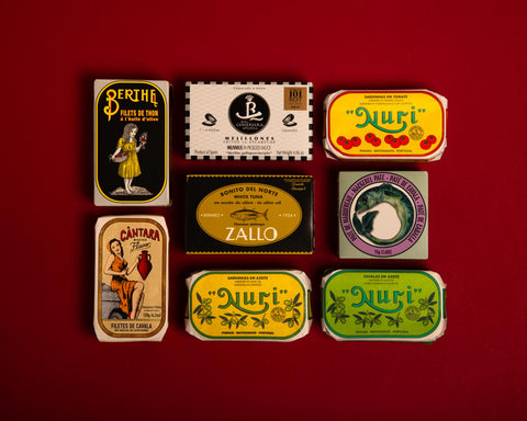 Eight tins on a red table top. The tins have colourful wrappers, one is yellow and is decorated with illustrations of tomatoes. Another tin has an illustration of a Portuguese lady holding a terracotta vase.