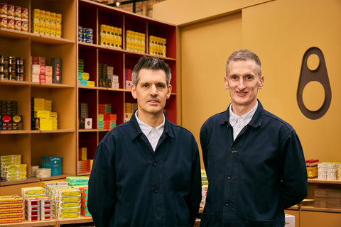 A photo of Patrick - the founder of the company, and Stephen - the store manager at Borough Market, standing in front of the colourful tins on display at the stall, wearing navy blue shirts.