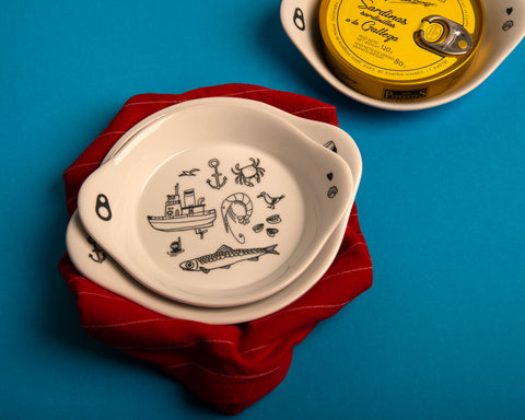 A stack of white plates on a red tablecloth, with an additional plate displaying a round golden tin of sardines positioned in the top, righthand corner.