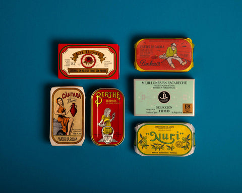 A selection of tinned fish arranged against a blue backdrop, featuring a yellow tin from the renowned Portuguese cannery Nuri.