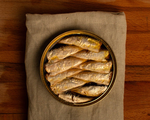 An open circular tin of hand-arranged small sardines on a grey cloth against a wood background