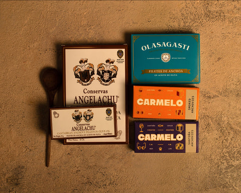 A selection of five tins of Cantabrian anchovies against a beige background, with a wooden spoon to the left of the tins