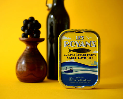 An upright tin of sardines in front of a glass bottle and wooden vase with wooden grapes on top.