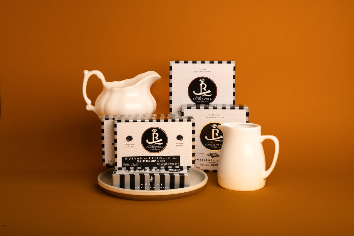 A selection of differently sized tins of Galician fish and shellfish in Real Conservera's trademark black and white packaging , with a white porcelain milk jug and cream jug and a grey and white plate, against a yellow background