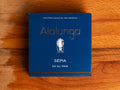 A tin of cuttlefish in ink in square-shaped blue card packaging  against a wood background. The packaging has Alalunga written in silver lettering above a white illustration of a cuttlefish.