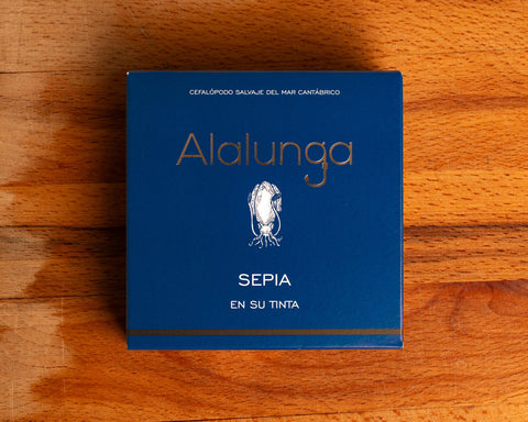 A tin of cuttlefish in ink in square-shaped blue card packaging  against a wood background. The packaging has Alalunga written in silver lettering above a white illustration of a cuttlefish.