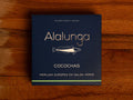 A tin of hake cheeks in green sauce, in square, blue, card packaging with Alalunga written in silver lettering above an illustration of a hake.