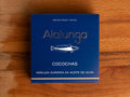 A tin of hake cheeks in olive oil in square-shaped blue card packaging  against a wood background. The packaging has Alalunga written in silver lettering above a white illustration of a hake.