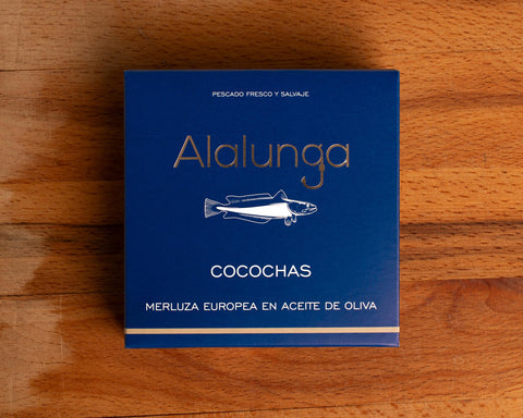 A tin of hake cheeks in olive oil in square-shaped blue card packaging  against a wood background. The packaging has Alalunga written in silver lettering above a white illustration of a hake.