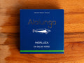 A tin of hake in salsa verde in blue card packaging  against a wood background. The packaging has Alalunga written in silver lettering above a white illustration of a hake, and a green strip along the bottom.