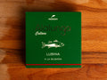 A tin of sea bass a la bilbaina or Bilbao style in green card packaging  against a wood background. The packaging has Alalunga written in silver lettering above a white illustration of a sea bass, and a red strip along the bottom.