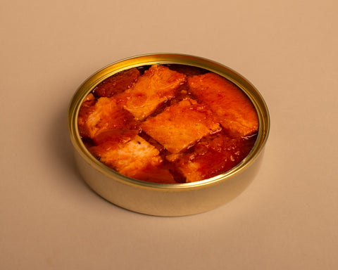 An open, circular, golden-coloured  tin with tuna fillets in tomato sauce, on a pale background. 
