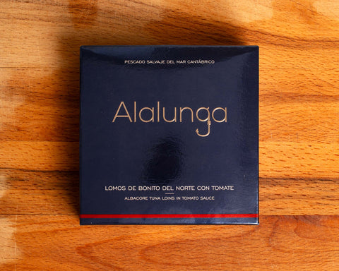 Canned white tuna in square blue card packaging with Alalunga in silver, embossed lettering above a diagram of a tuna. There is a red stripe along the bottom of the packaging.