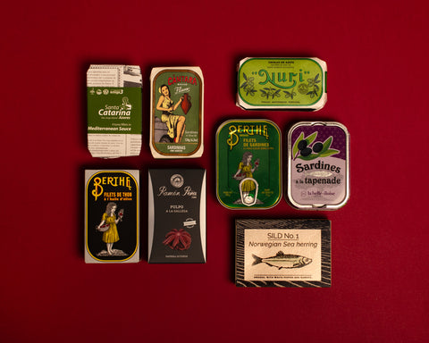 A selection of colourfully-packaged tinned tuna, sardines, mackerel, herring and octopus against a red background.