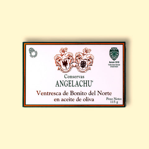 A tin of ventresca or tuna belly in a rectangular card packaging. The box is white with a dark green and golden border. Conservas Angelachu is written in dark green lettering and ventresca de Bonito del Norte en aceite de oliva in gold leaf lettering, beneath a coat of arms. The box is against a yellow wood background.