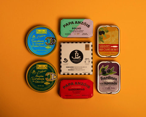 Collection of premium tins on an orange table. There are sardines, octopus and mussels from Spain and France