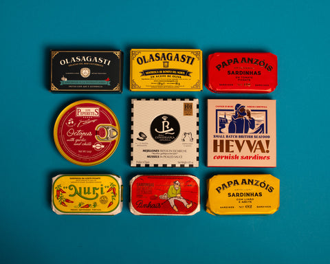 A selection of colourfully-packaged tinned sardines, tuna belly, octopus and mussels  against a blue background.