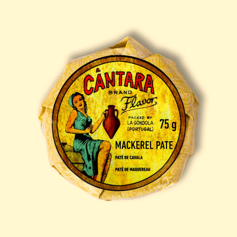 A round tin of mackerel pâté in a glossy paper wrap with an image of a woman in a green dress holding an amphora of olive oil on the front