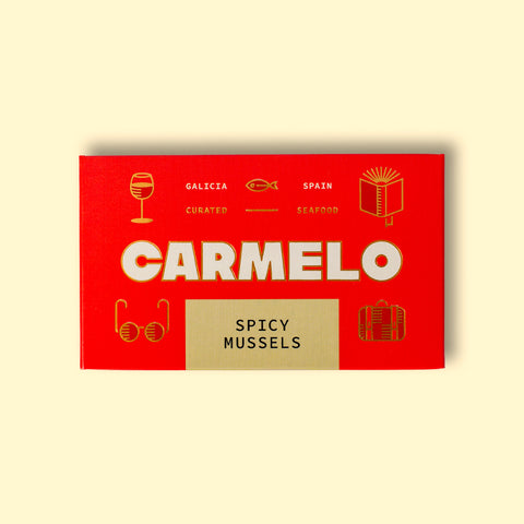 Red box of Carmelo Spicy Mussels on a wooden surface. The box is bright and has four small illustrations, a glass of wine, a book, a suitcase and a pair of glasses.