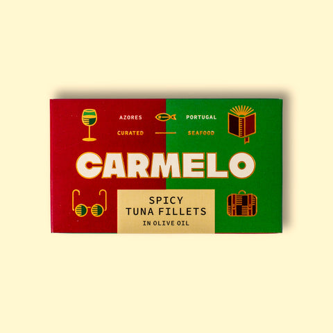 Carmelo spicy tuna fillets on a yellow wooden table. The paper box is green and red with big letters saying Carmelo and there are golden illustrations around the box