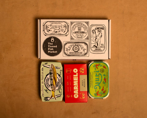 White illustrated box with three tins of fish. Two of them are wrapped in colourful green and pale green papers and the other comes in a red luxurious box.