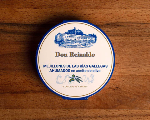 A round tin with a blue and white card wrap. There is an olive branch at the base, above it Don Reinaldo is written in gold lettering and there is a drawing of the cannery.