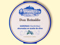 A round tin with a white card wrap. The cover has a blue and gold border. Don Reinaldo is written in gold lettering below a drawing of the cannery.