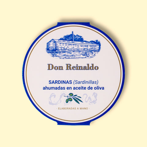 A round tin with a white card wrap. The cover has a blue and gold border. Don Reinaldo is written in gold lettering below a drawing of the cannery.