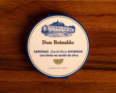 A round tin in white with blue borders featuring the brand name Don Reinaldo in brown lettering and an illustration of a cannery above with a lemon slice image below. 