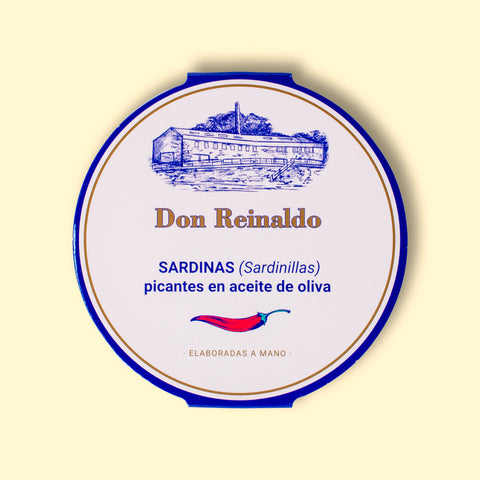 A round tin with a white card wrap. The cover has a blue and gold border. Don Reinaldo is written in gold lettering below a drawing of the cannery. and above a red chilli pepper.