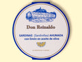 A round tin in white with blue borders featuring the brand name Don Reinaldo in brown lettering and an illustration of a cannery above with a lemon slice image below.