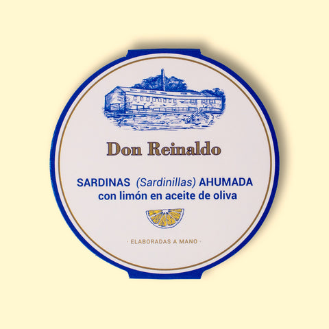 A round tin in white with blue borders featuring the brand name Don Reinaldo in brown lettering and an illustration of a cannery above with a lemon slice image below. 